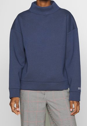 Granatowy sweter z wysokim kołnierzem, opadającymi ramionami i ściągaczami. Posiada subtelne logo na rękawie oraz miękką, teksturowaną tkaninę.