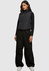 Svart puffer-vest med høy krage og quiltet mønster, kombinert med løstsittende svarte bukser og hvite sneakers.