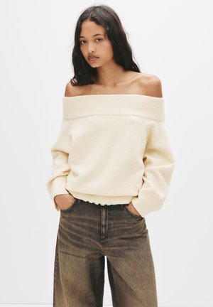 Maglione crema off-the-shoulder con scollatura a costine e polsini, abbinato a jeans marrone chiaro a vita alta con una leggera texture.
