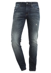 Vaqueros de denim en azul oscuro con un lavado desgastado. Presentan parches desgastados, diseño estándar de cinco bolsillos y una cremallera con cierre de botón.