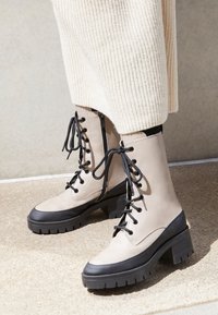 Bottes de cheville en cuir beige avec semelles en caoutchouc noires, à lacets à l'avant et fermeture éclair sur le côté. Présente un dessus texturé et des accents noirs contrastants.