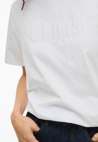 Hvid bomulds t-shirt med broderet tekst "CHÉRIE" i midten. Kombineret med mørkeblå denimjeans. Glat tekstur, afslappet pasform.