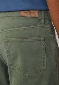 Maslinasto zelene pamučne hlače s glatkom teksturom. Imaju svijetlosmeđi kožni detalj s utisnutim natpisom "HACKETT" i stražnji džep s detaljima šivanja.