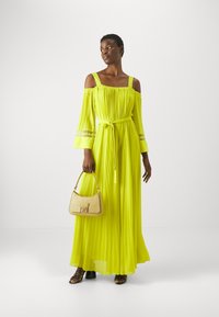 Longo vestito off-shoulder in tessuto plissettato giallo brillante con vita adatta, abbinato a una piccola borsa beige e sandali neri con tacco.