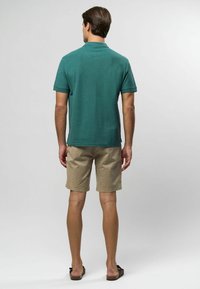 Tmavofialová polokošeľa s krátkymi rukávmi, príjemná na dotyk, nosená s béžovými šortkami, zobrazená zozadu na svetlom pozadí.