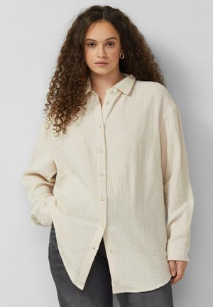 Femme aux cheveux longs et bouclés portant une chemise crème texturée à boutons et un pantalon foncé, debout devant un fond clair uni.