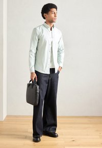 Ανοιχτό πράσινο πουκάμισο με κουμπιά, σκούρα τζιν με φαρδιές παντελόνες, μαύρα παπούτσια, κρατώντας μια συμπαγή μαύρη τσάντα με λαβές στο πάνω μέρος. Απλός, σύγχρονος σχεδιασμός.