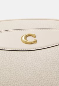 Světle béžová kožená kabelka s texturovaným povrchem, s akcentem z pozlaceného monogramovaného kovu a prošívaným lemem v horní části.