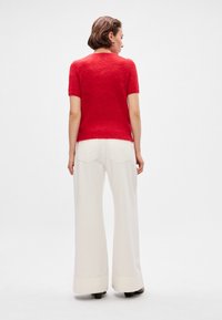 Piros, rövid ujjú pulóver, puha, textúrált anyagból, széles lábú, krémszínű nadrággal párosítva, amelynek enyhe harangformája van, és hátsó zsebei is vannak.