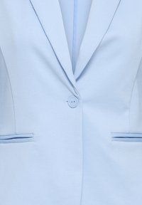 Blazer bleu clair, tissu texturé, fermeture à un bouton, avec deux poches latérales décorées d'un liseré contrastant, design à revers.