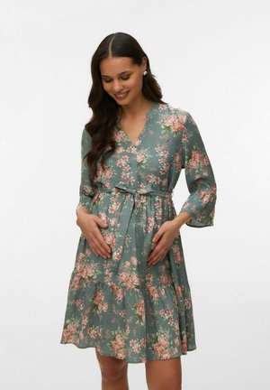 Femme enceinte souriante portant une robe verte à fleurs jusqu'aux genoux avec des manches trois-quarts, les mains posées sur son ventre.