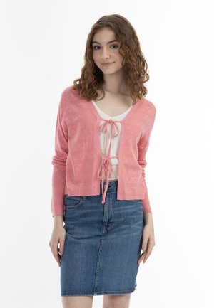Junge Frau mit lockigem Haar, die eine pinke Strickjacke trägt, vorne gebunden, über einem weißen Top und einem blauen Jeansrock.