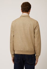 Chaqueta bomber beige con una tela texturizada, puños y dobladillo de canalé, y un corte relajado, combinada con unos jeans azul oscuro que cuentan con bolsillos traseros.