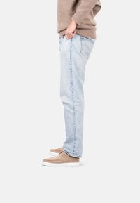 Jeans de mezclilla azul claro de corte recto, combinados con unos zapatos slip-on de ante color marrón claro. El modelo lleva un suéter marrón, de pie de lado.