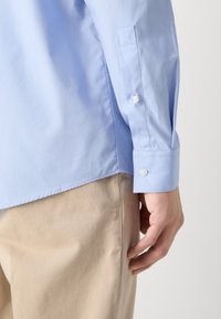 Homme portant une chemise bleu clair à manches longues et un pantalon beige, mettant l’accent sur le poignet de la manche avec des boutons et la main reposant sur le côté.