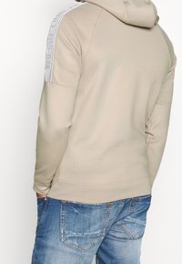 Beige hoodie med en figurnära design, med en texturerad axeldetalj med varumärke och ett mjukt, slätt material. Bärs tillsammans med blekta blå jeans.