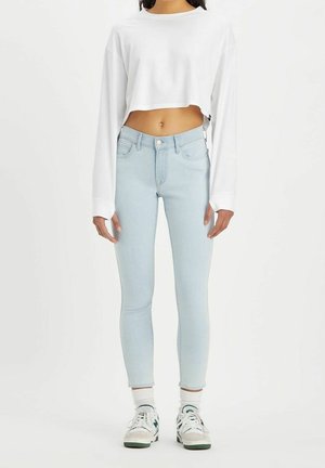 Personne portant un jean skinny bleu clair, un t-shirt blanc court à manches longues, des chaussettes blanches et des baskets blanches avec des accents vert foncé, debout.