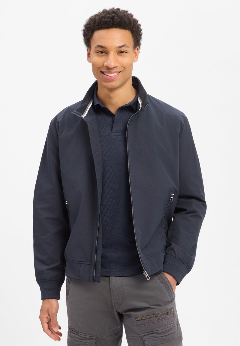 Lächelnder Mann, der eine marineblaue Jacke mit Reißverschluss über einem dunklen Poloshirt und grauen Cargo-Hosen mit Reißverschlusstaschen trägt, steht mit einer Hand in der Tasche.