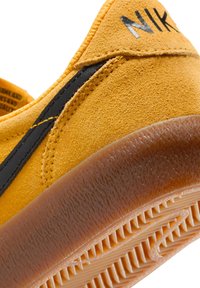 Sneaker giallo in suede con accenti neri, dotato di suola in gomma testurizzata e logo prominente. Primo piano del lato e della suola.