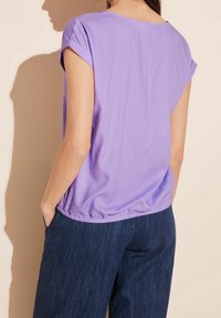 Haut lavande à manches courtes avec un ourlet froncé, fabriqué en tissu léger. Porté avec un pantalon en denim noir, montré de dos.