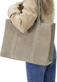 Grijze suède shoppers tas met een gestructureerd ontwerp, voorzien van twee verticale naden en lange handvatten, gedragen op een schouder met een crème-kleurige top.