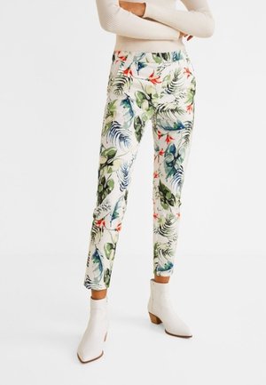 Person iført hvide ankelstøvler og tætsiddende bukser med farverigt tropisk blad- og blomstermønster, stående med armene krydset.