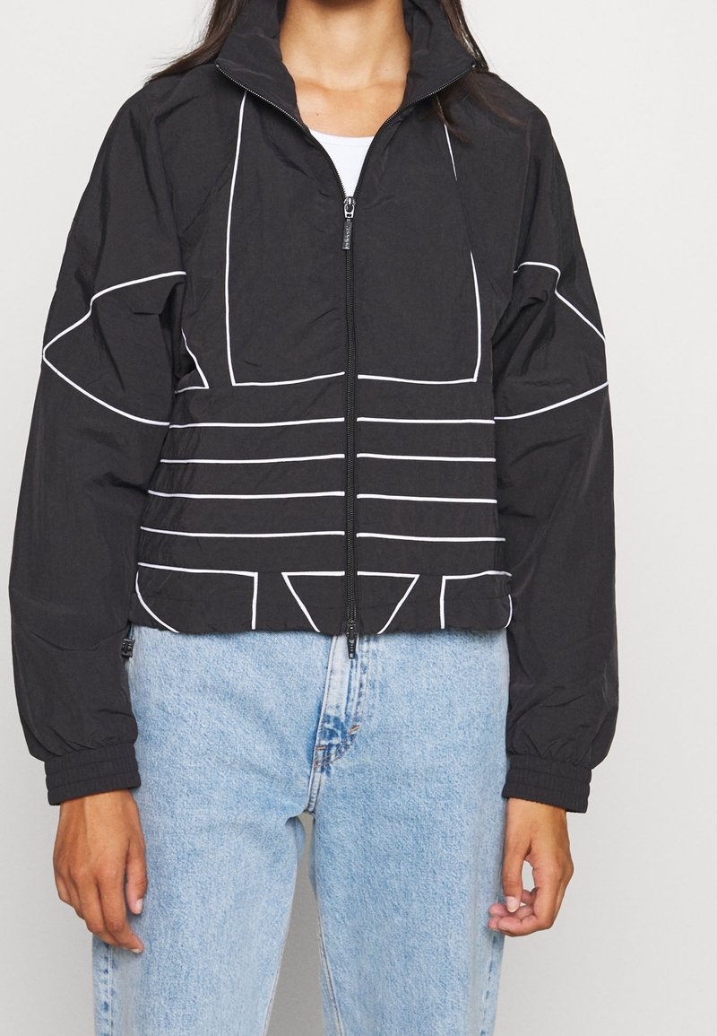 Svart kort jacka med vita geometriska linjer, dragkedjeöppning, hög krage och elastiska mudd. Sammansatt med ljusblå denimjeans.
