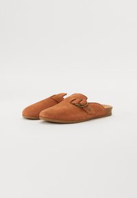 Brown suede slip-on shoes with an open back, featuring a side buckle detail and a textured cork insole. Flat rubber sole.