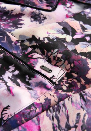 Lähedalt vaade lillemustriga kangale, mis on lilase, roosa, musta ja kreemja tooni kombineerimisega, millel on tikitud tasku ja väike metallist silt, millel on kirjutatud "NEXT."