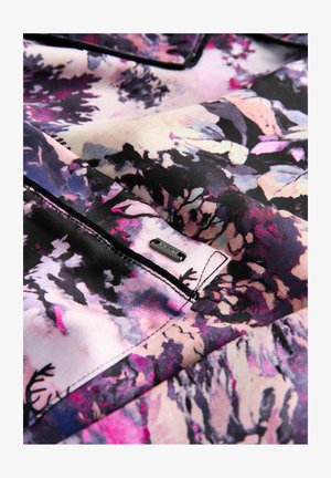 Artimas florų raštuotos medžiagos nuotrauka, kurioje dominuoja purpurinė, rožinė, juoda ir kreminė spalvos, su siuvinėta kišenėle ir mažyte metaline etiketėle su užrašu "NEXT."