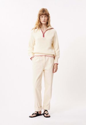 Pull crème avec fermeture éclair quart de zip et bordure rose, associé à un pantalon beige clair et des sandales noires. Coupe décontractée et tissu texturé.