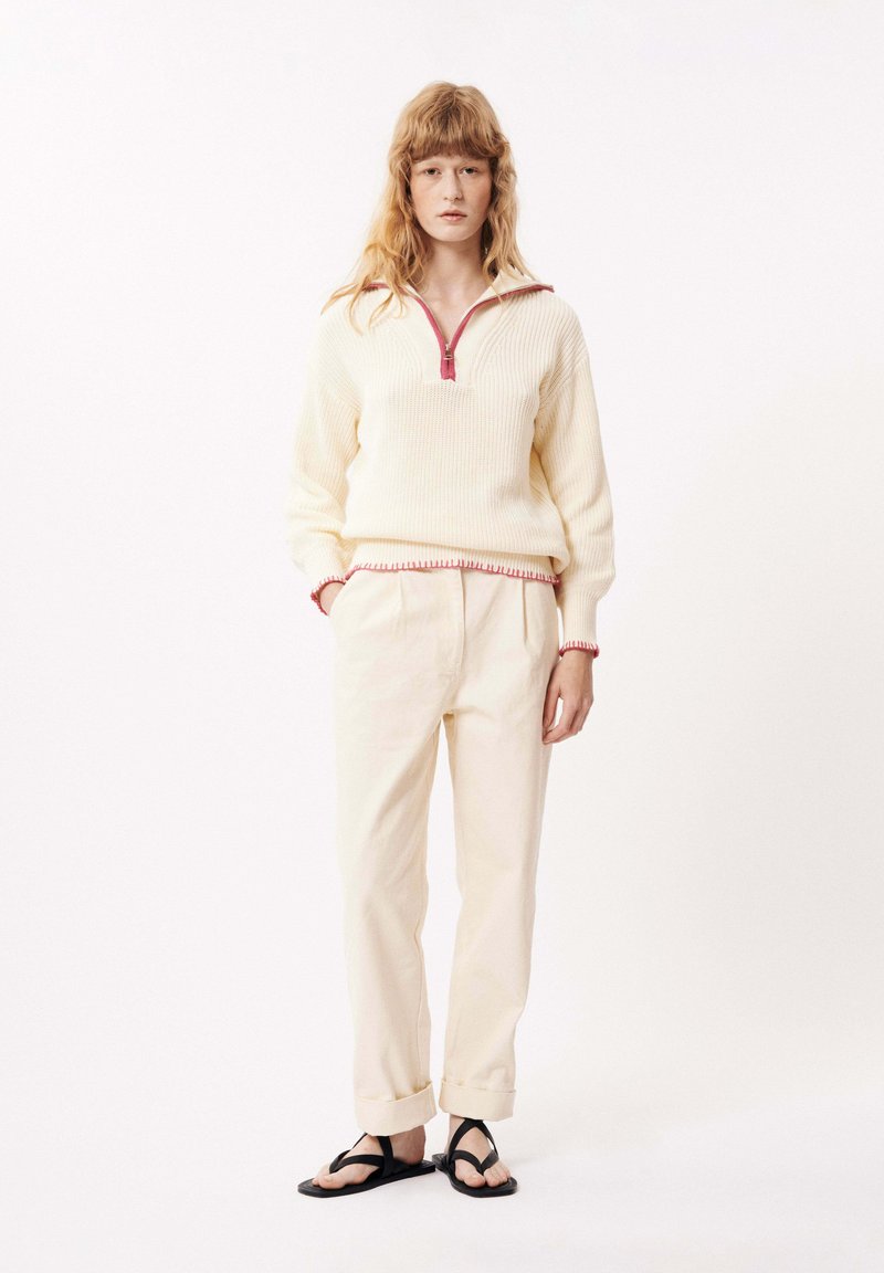 Pull crème avec fermeture éclair quart de zip et bordure rose, associé à un pantalon beige clair et des sandales noires. Coupe décontractée et tissu texturé.