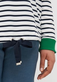 Top a righe bianche e blu navy con polsini a coste verdi, dotato di cordino con occhielli in argento. Abbinato a pantaloni blu.