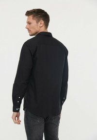 Chemise noire à manches longues en coton, dotée d'un col à boutons, de fentes latérales et de boutons aux poignets, présentée de dos.