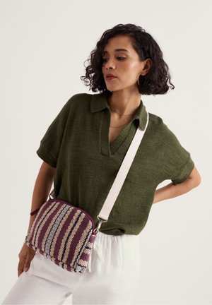 Femeie care poartă un top tricotat cu mâneci scurte de culoare verde măslinie și pantaloni albi, având o geantă crossbody cu dungi mov, bej și albastre, cu o curea largă albă.