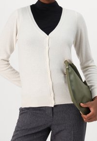 Žena v krémovém zapínacím kardiganu přes černý rolák, drží zelenou kabelku a má na sobě tmavé kalhoty s herringbone vzorem.