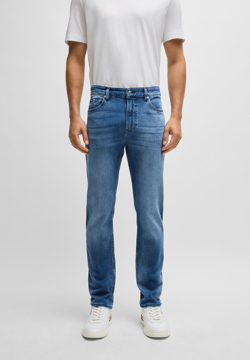 Homme portant un jean slim bleu clair, des baskets blanches et un t-shirt blanc uni à manches courtes, debout devant un fond gris.