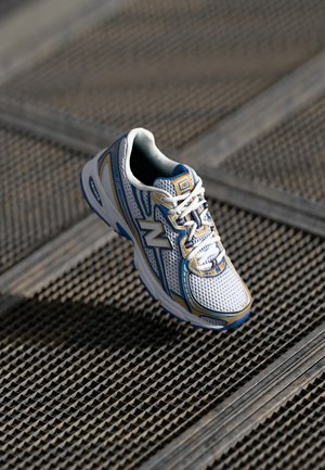 Witte en blauwe New Balance hardloopschoen met veters op een metalen roosteroppervlak met een getextureerd roosterpatroon.