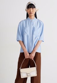 Light blue short-sleeve shirt with a high collar and two front pockets, paired with brown wide-leg trousers and a white handbag with a metal accent.