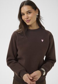 Rjav bombažni pulover z dolgimi rokavi, rebrastimi manšetami in okroglim izrezom. Na prsih majhen rožnat logotip v obliki srca.