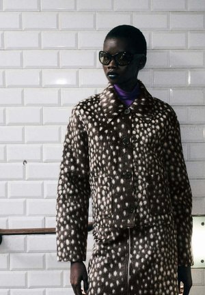 Persona con chaqueta y falda marrón y blanca con manchas y gafas de sol oscuras, de pie contra una pared blanca con azulejos.