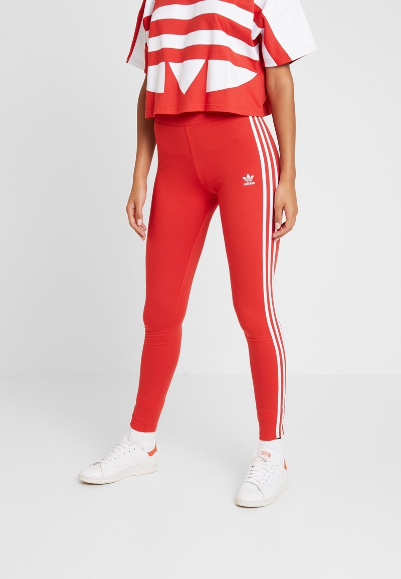 Röda leggings i elastisk tyg med vita sidostreck och en adidas-logotyp nära midjan, tillsammans med en röd och vit randig crop top.