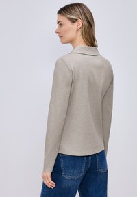 Femme aux cheveux blonds raides portant un t-shirt à manches longues gris clair et un jean bleu, debout de profil gauche, dos visible.