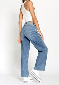 Žena v tílku bez rukávů, bílých pletených topu, modrých džínech s vysokým pasem a bílých platformových teniskách, pózující z boku na jednoduchém pozadí.