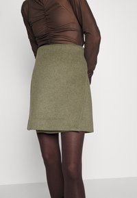Olijfgroene wollen minirok met een rechte snit en zijnaad, gecombineerd met donkere semi-doorzichtige panty's. Getextureerde, warme stof.