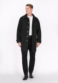 Cappotto lungo nero con colletto, bottoni argentati e tasche laterali, abbinato a una camicia bianca e pantaloni neri, su uno sfondo bianco.