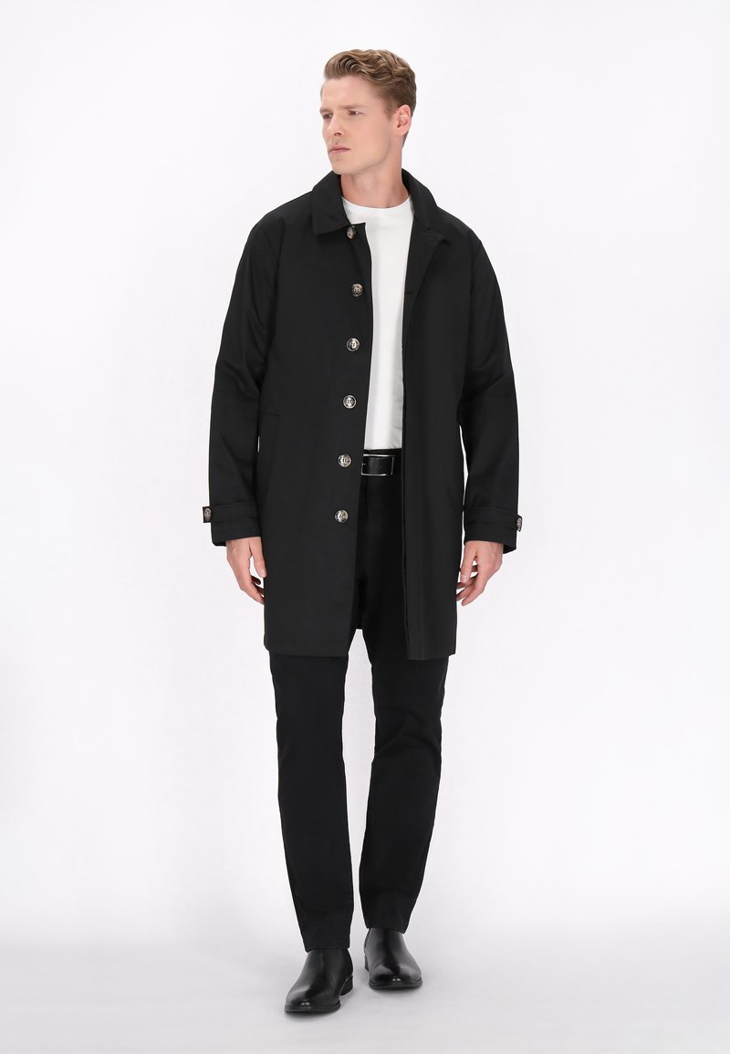 Cappotto lungo nero con colletto, bottoni argentati e tasche laterali, abbinato a una camicia bianca e pantaloni neri, su uno sfondo bianco.