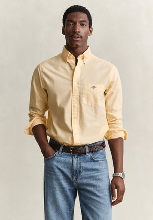 Homme aux cheveux courts et bouclés avec moustache, portant une chemise jaune boutonnée avec manches retroussées, jean bleu, ceinture marron et montre-bracelet argentée.