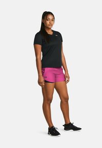 Camiseta atlética negra de manga corta, pantalones cortos rosas con acentos negros y zapatillas negras. Modelo en una pose de pie contra un fondo liso.