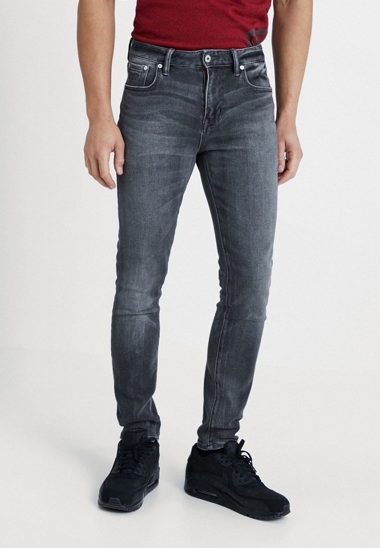 Hombre vistiendo jeans negros ajustados y desgastados, zapatillas negras y una camisa roja, de pie contra un fondo blanco.
